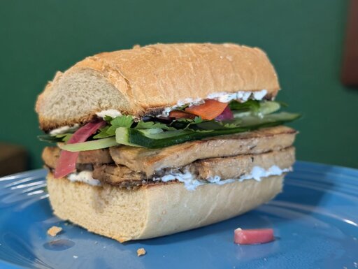 A sandwich on a blue plate, made from a toasted French roll, with layers of tofu, colorful vegetables, and a yogurt sauce visible.
