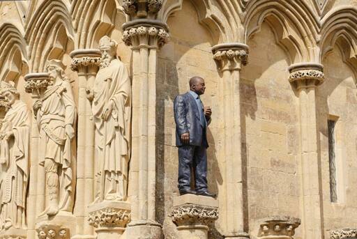 A modern painted sculpture of a man in a suit holding a takeout coffee cup installed alongside early English Gothic sculptures