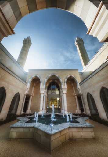 A symmetrical courtyard view of the Masjid e Vali mosque in Blackburn, photographed from ground level looking upwards through an open-air atrium, revealing pale sandstone walls with three large horseshoe arches framing the entrance portal, ornamental white columns supporting the archways, and a central star-shaped fountain with cascading water jets in pale marble, all framed by the geometric angles of the surrounding cream-coloured arcading that leads the eye upward past two towering cylindrical minarets with their distinctive ribbed upper sections against a clear blue sky with wispy clouds.